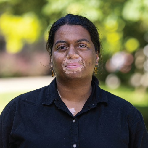 Subhanya Sivajothy with black hair, black button-up shirt and a serious expression stands outdoors on campus.