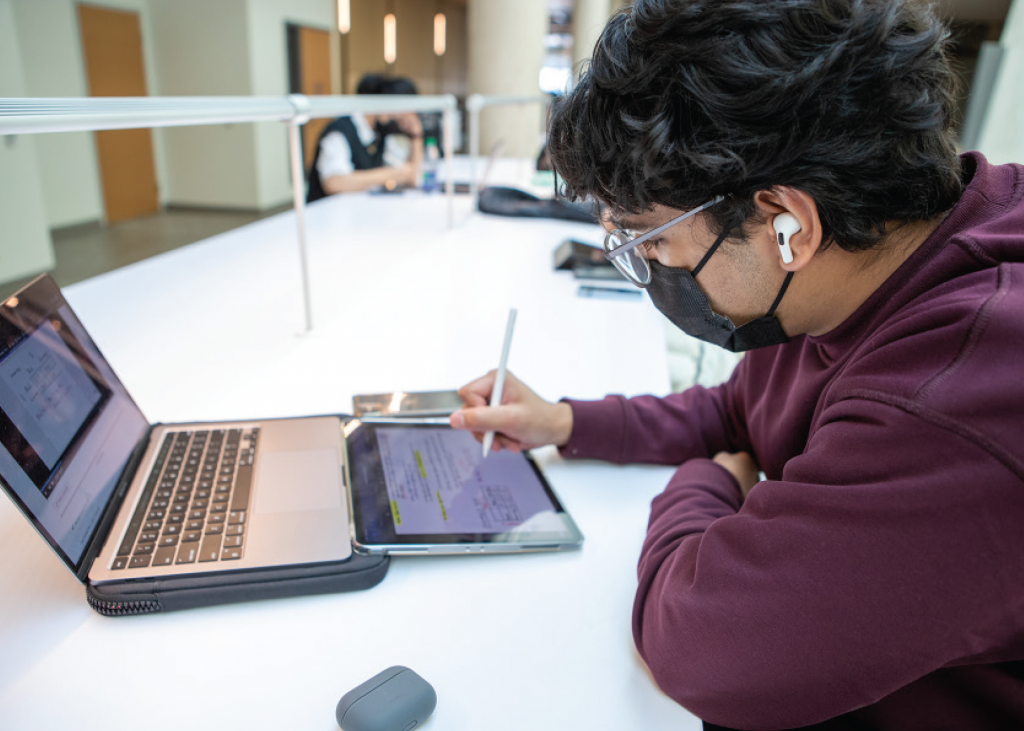 A student with dark hair, glasses, ear buds, maroon sweater and focused expression sits at a laptop taking notes.