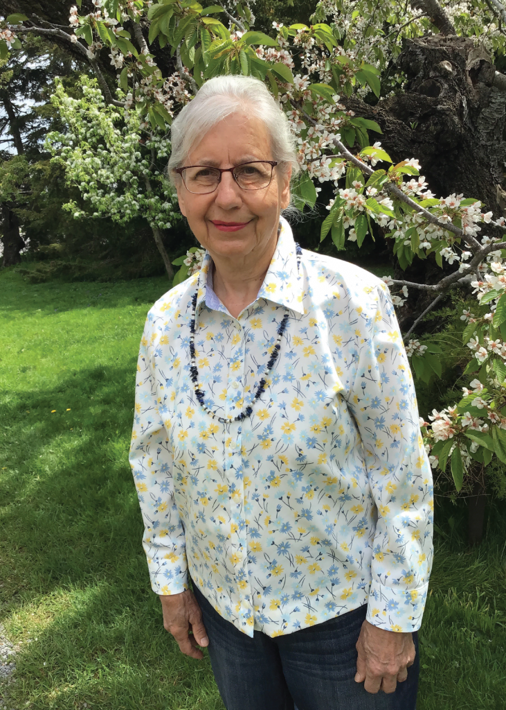 Carol Mazur with silver hair, glasses and a floral blouse, stands smiling outdoors by a blossoming tree.