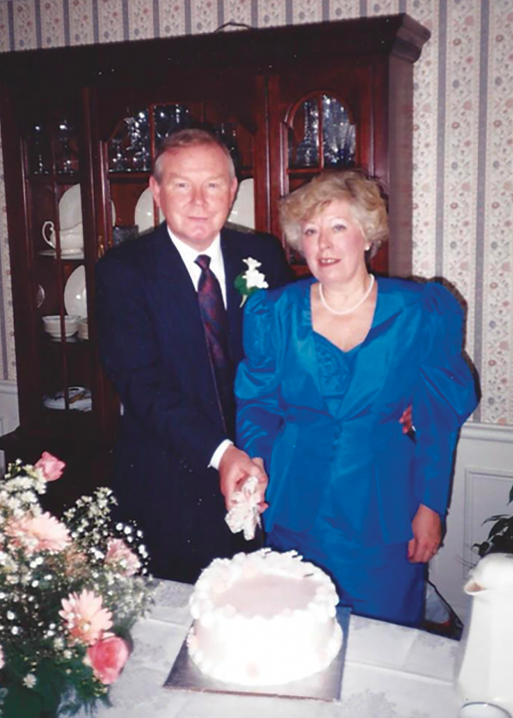 Vic Nunn dressed in a black suit and Carol Nunn wearing a blue dress cutting a piece of celebration cake together in a home.
