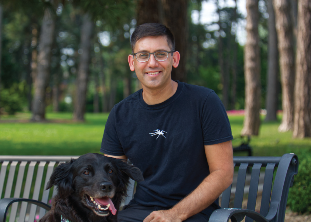 Pasha Malla with black hair, glasses, black T-shirt and a smile, sits on a park bench with a black Labrador retriever.