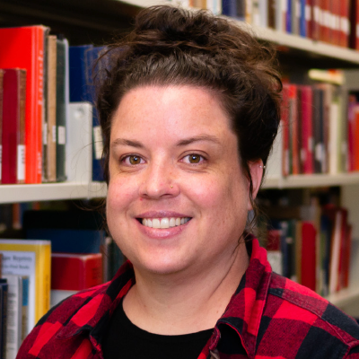 Bronwen Glover with black hair tied up, and a red and black plaid shirt over a black shirt, standing in front of a library bookshelf.