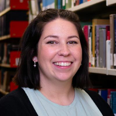 Carlie King with shoulder-length black hair and a blue and black shirt, standing in front of a library bookshelf.