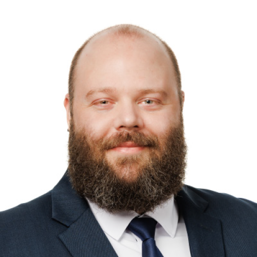 Casey Hoeve with short brown hair, brown beard and navy suit and tie sitting in front of a white background.