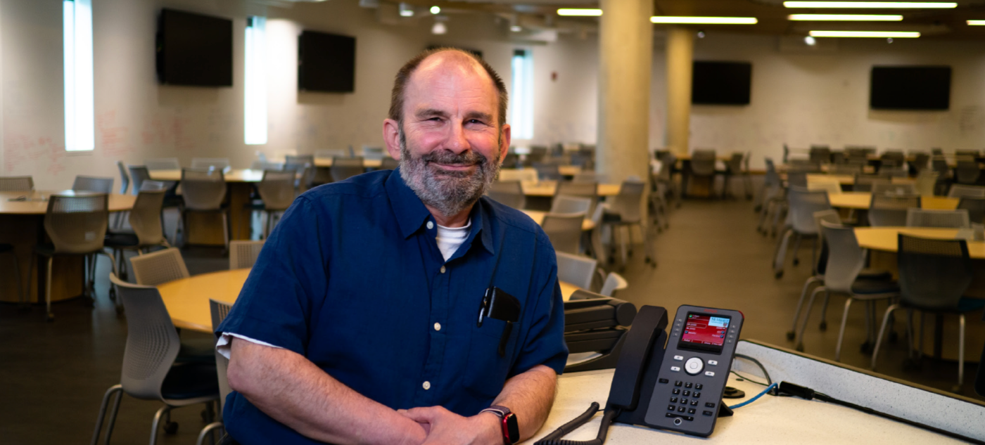Kevin Andrews with short brown hair, grey beard, and blue jean shirt stands at a desk with a black phone inside an empty university classroom. 