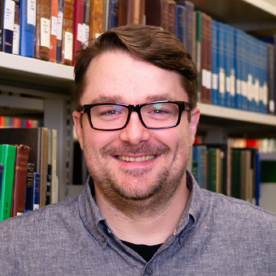 Matthew Burns with short brown hair, glasses and a blue dress shirt, standing in front of a library bookshelf.