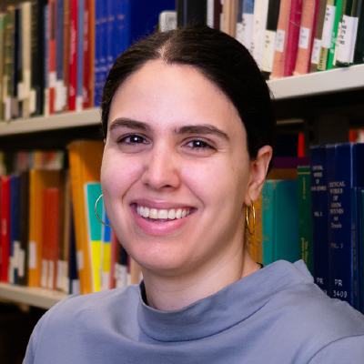 Saman Goudarzi with black hair tied back and a blue blouse, standing in front of a library bookshelf.