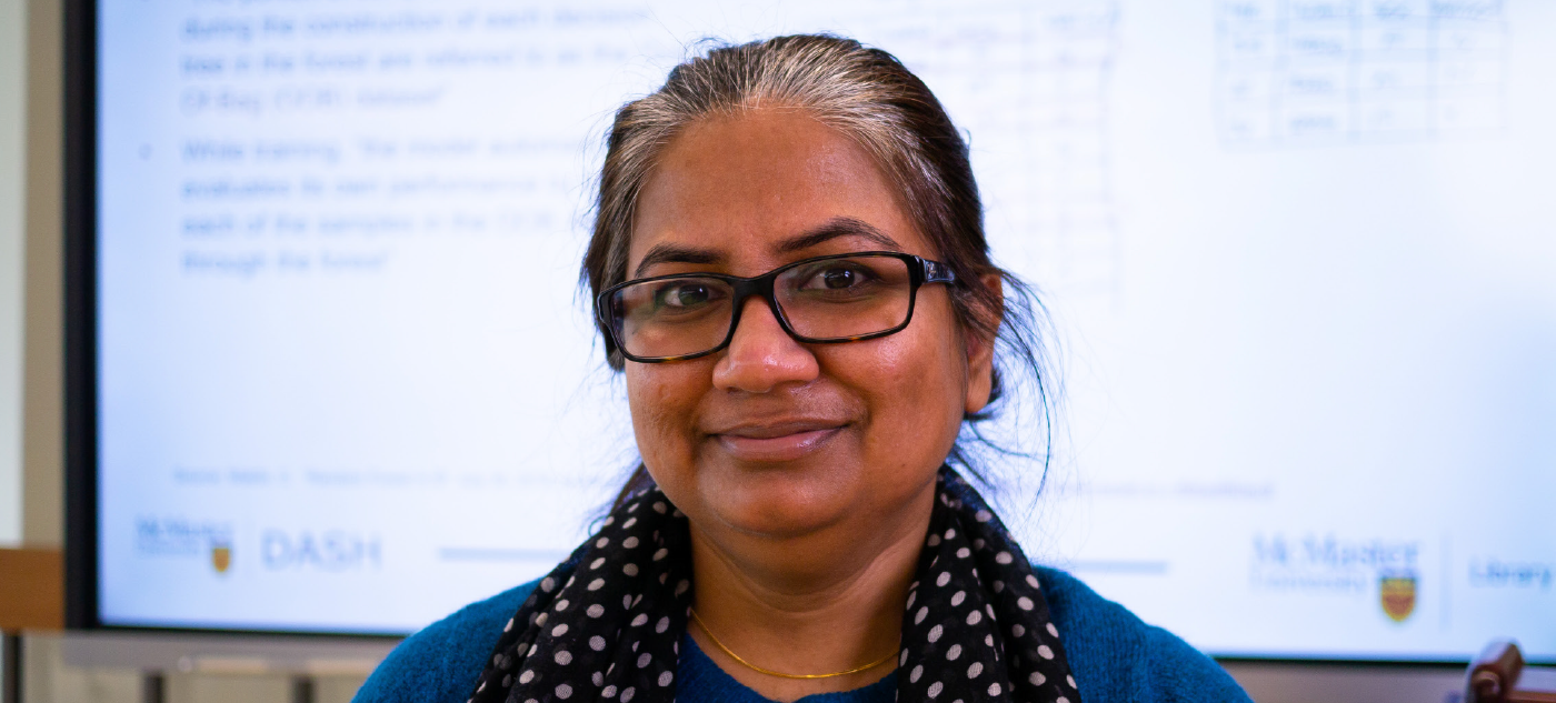Shaila Jamal with grey-black hair pulled back, glasses and a polka-dot scarf over a blue sweater, in front of a digital screen in a classroom. 