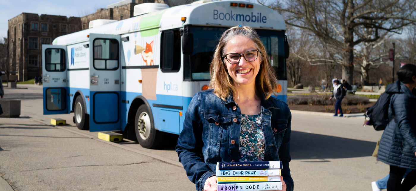 Lynne Serviss with shoulder-length brown hair wearing a jean jacket smiling and standing holding books in front of the Hamilton Public Library bookmobile outside on a sunny day.