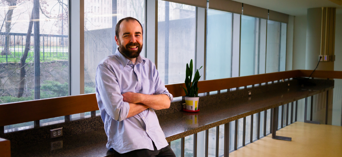 Jack Young with short brown hair and beard wearing a blue dress shirt smiling with arms crossed leaning against a counter in front of a window.