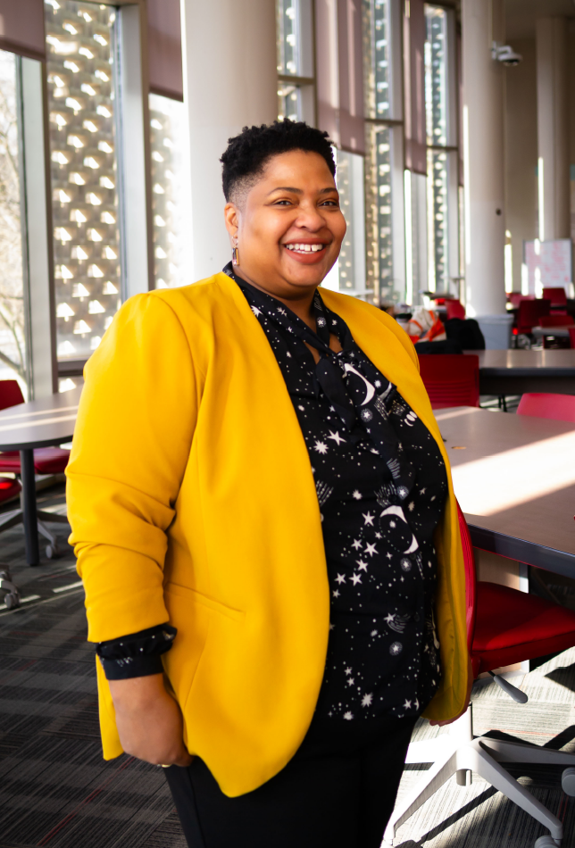 Crystal Mark with short black hair wearing a yellow jacket over a black blouse smiling in front of a window in Mills Learning Commons.