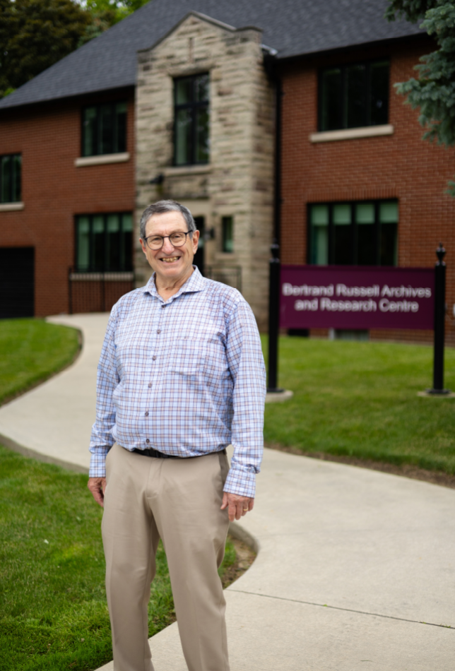 Bernard Linksy with short grey hair and glasses standing and smiling outside in front of a brick two-storey building.