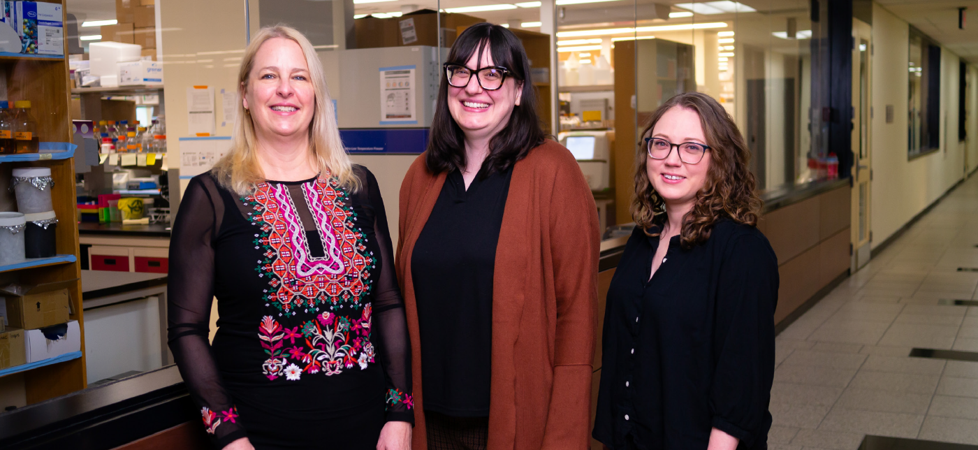 Joanne Kehoe with long blonde hair, Felicia Vulcu with long brown hair, and Katie Harding with curly brown hair smiling and standing in a hallway in front of a laboratory.