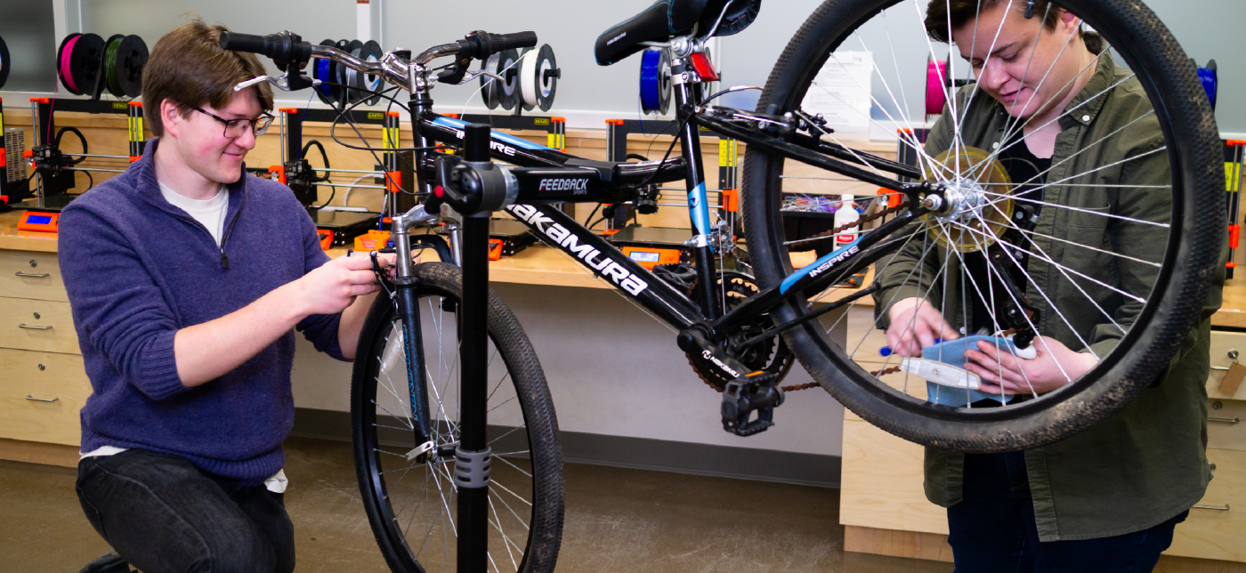 Vlad Brundula-Ilija with short brown hair and kneeling, and Brittany Sostar with short brown hair and standing, repairing a bicycle inside a workspace with 3D printers in the background